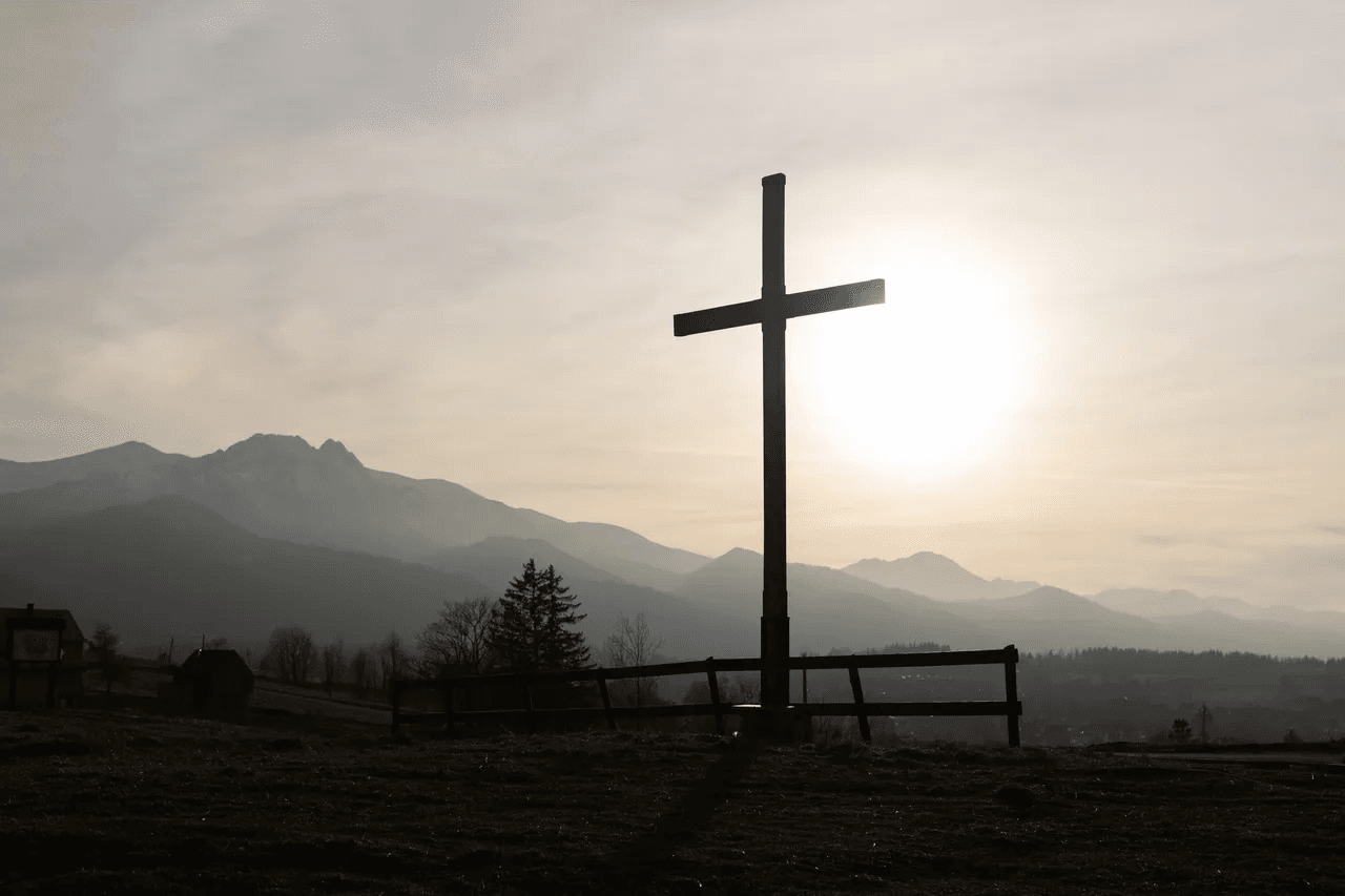 A wooden cross silhouetted against the sun above a hillside fence, with mountains in the distance